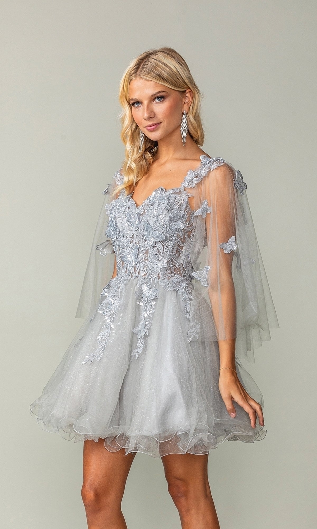 A woman wears the Butterfly-Cape Short A-Line Homecoming Dress 3393 in silver tulle with floral appliqué and sheer sleeves, standing against a plain background.